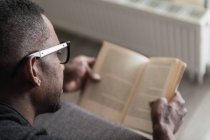 Крупным планом афроамериканец в очках читает книгу, расслабляясь дома на диване — стоковое фото