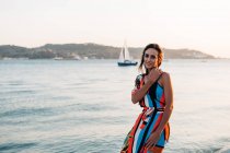 Вміст жінки в довгій сукні, що стоїть на узбережжі на заході сонця проти морського пейзажу — стокове фото
