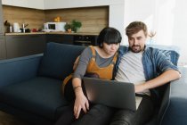 Молодой человек и женщина обнимают друг друга и наслаждаются хорошей пленкой на ноутбуке, сидя на удобном диване в уютной гостиной — стоковое фото