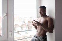 Без сорочки м'язистий чорний чоловік використовує мобільний телефон, стоячи проти вікна в денне світло — стокове фото