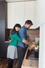 Side view of beautiful young female keeping eyes closed and hugging cooking man from back while standing in stylish kitchen together — Stock Photo