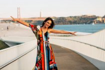 Елегантна жінка в одязі, що стоїть на мосту з руками, витягнутими на сонячному світлі — стокове фото