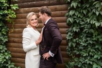 Eleganti sposi adulti che si abbracciano in piedi in giacca e cravatta contro la parete di legno e sorridono alla macchina fotografica — Foto stock