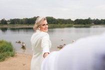 Erwachsene schöne und elegante Braut hält die Hand des Bräutigams und lächelt in die Kamera, während sie mit Schwänen am Seeufer steht — Stockfoto