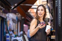 Женщина с напитком и смартфоном возле открытого кафе — стоковое фото
