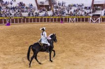Spain, Tomelloso - 28. 08. 2018. View of female bullfighter riding horse on sandy area with people on tribune — Stock Photo