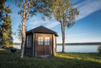 Сцена маленького дерев'яного будинку, розміщеного на узбережжі синього ще озера між деревами на тлі чистого неба — стокове фото