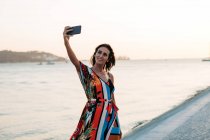 Mujer contenta en colorido vestido de verano usando el teléfono y tomando selfie en primera línea de mar al atardecer - foto de stock
