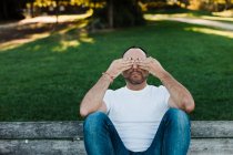 Привабливий дорослий чоловік прикриває очі, сидячи на лавці в парку в сонячний день — стокове фото