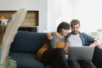 Young man and woman embracing each other and enjoying good film on laptop while sitting on comfortable sofa in cozy living room — Stock Photo