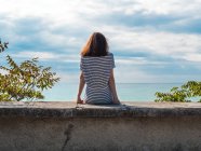 Femme voyageuse assise sur une clôture en pierre sur fond de ciel nuageux et de paysage marin bleu — Photo de stock