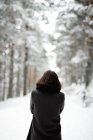 Красивая молодая женщина в стильном наряде смотрит в сторону, стоя рядом с деревьями, покрытыми снегом в холодный день в прекрасной сельской местности — стоковое фото