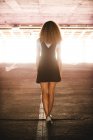 Vue arrière de la mince femme bouclée portant une robe noire avec des baskets debout sur un parking souterrain à la lumière du soleil — Photo de stock