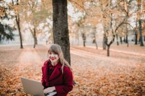 Jovem senhora de casaco vermelho usando dispositivo e sentado no assento na floresta de outono — Fotografia de Stock