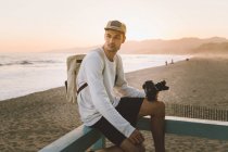 Vista lateral de cara atraente com mochila e câmera de foto profissional sentado no corrimão na praia de areia e olhando para longe durante o belo pôr do sol em Santa Monica, Califórnia — Fotografia de Stock