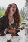Lovely young female holding cute cat and laughing while sitting at table near countryside building in Bulgaria, Balkans — Stock Photo