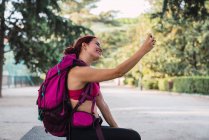 Mujer joven sonriente en ropa deportiva con mochila rosa sentada en el parque y tomando selfie con teléfono inteligente - foto de stock