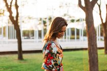 Elegante donna bruna sorridente in abito colorato in piedi nel parco urbano e guardando in basso — Foto stock