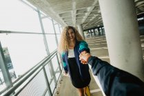 Schöne junge Frau mit lockigem Haar lächelt und hält die Hand eines anonymen Freundes, während sie anbietet, ihr auf einem überdachten Weg zu folgen — Stockfoto