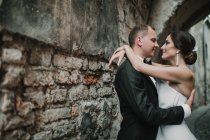 Vista laterale di attraente giovane sposa e sposo che si abbracciano e si guardano mentre si trovano vicino al muro crollante di un antico edificio sulla strada della città — Foto stock