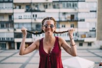 Cheerful red-haired young woman in sunglasses holding braids against residential building — Stock Photo