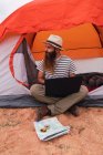 Attraente ragazzo barbuto utilizzando il computer portatile e guardando lontano mentre seduto vicino tenda e mappa con bussola — Foto stock