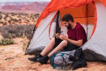 Barbudo cara em roupa casual segurando caneca de bebida quente e smartphone moderno enquanto sentado perto da tenda e olhando para longe na bela natureza — Fotografia de Stock