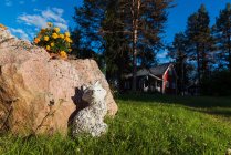 Statue of dog made of white stone near big rock with flowers with wooden house among trees on background — Stock Photo