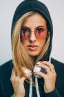 Young blond woman in black hoodie and round sunglasses with headphones on neck looking at camera on white background — Stock Photo