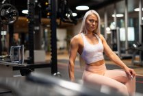 Jeune femme blonde en vêtements de sport assis dans la salle de gym et regardant la caméra sur le fond de l'équipement — Photo de stock
