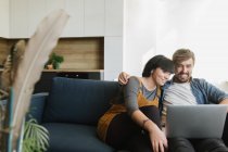 Young man and woman embracing each other and enjoying good film on laptop while sitting on comfortable sofa in cozy living room — Stock Photo