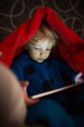 Menino em pijama usando tablet digital sob cobertor — Fotografia de Stock