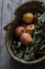 Frische reife Äpfel und Blätter im Korb auf Holztisch — Stockfoto