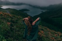 Rückansicht einer jungen Frau, die auf einem Hügel mit grünem Gras steht und auf den schönen See unten blickt — Stockfoto