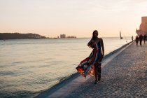 Вміст жінки в довгій барвистій сукні, що йде на кам'яній набережній на заході сонця проти морського пейзажу — стокове фото