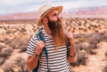 Guapo barbudo con mochila mirando hacia otro lado mientras está de pie sobre un fondo borroso del desierto increíble - foto de stock
