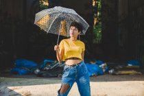 Jeune femme réfléchie en tenue élégante posant avec parapluie transparent tout en se tenant dans la rue le jour ensoleillé — Photo de stock