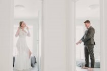 Belle jeune femme en robe blanche et beau garçon en costume élégant se préparant pour la cérémonie de mariage dans différentes chambres d'hôtel — Photo de stock