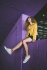 Loira encaracolado elegante em tênis e casaco amarelo sentado na parede roxa e rindo — Fotografia de Stock