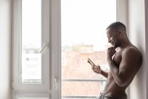 Sorrindo muscular homem negro usando telefone enquanto de pé contra a janela à luz do dia — Fotografia de Stock