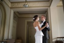 Вид сбоку привлекательной невесты и жениха, улыбающихся и смотрящих друг на друга, танцующих внутри красивого роскошного здания — стоковое фото