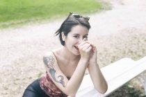Mulher sorridente atraente com cabelo escuro e tatuagem no ombro sentado no banco no parque e olhando para a câmera — Fotografia de Stock