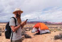 Maschio barbuto guardando lontano e navigando smartphone moderno mentre in piedi nel deserto vicino tenda e amico — Foto stock