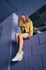 Elegante donna bionda riccia in scarpe da ginnastica e giacca gialla seduta sul muro viola e ridendo — Foto stock