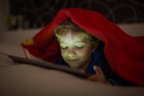 Усміхнений маленький хлопчик дивиться мультфільми з цифровим планшетом у ліжку під ковдрою — стокове фото