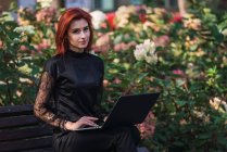 Retrato de jovem mulher elegante usando laptop no banco no parque — Fotografia de Stock