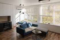 Удобный диван, стоящий рядом с кухонной мебелью в стильной комнате современной квартиры в солнечный день — стоковое фото