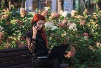 Jovem atraente com smartphone de navegação laptop enquanto sentado no banco no dia ensolarado — Fotografia de Stock