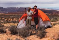 Два красивых мужчины стоят возле палатки и делают фотографии и видео удивительной пустыни, путешествуя вместе — стоковое фото