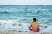 Vista trasera del macho en pantalones cortos sentados en la orilla arenosa cerca del magnífico mar ondulante en Tylenovo, Bulgaria - foto de stock